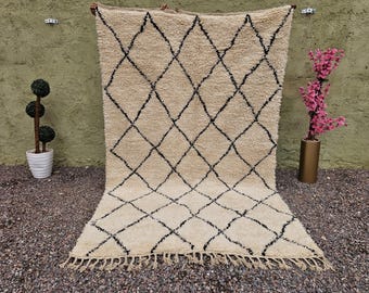 May include: A beige rug with a black diamond pattern. The rug has a shaggy texture and a fringe along the bottom edge. The rug is displayed against a green wall and a gravel surface.