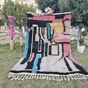 May include: A colorful abstract rug with geometric shapes in pink, blue, yellow, and black. The rug has a fringe border and is made of wool.