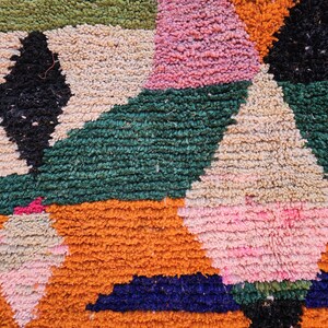 May include: Close-up of a colorful, textured rug with geometric patterns. The rug features a mix of colors including orange, green, pink, black, and cream. The design includes triangles and diamond shapes.