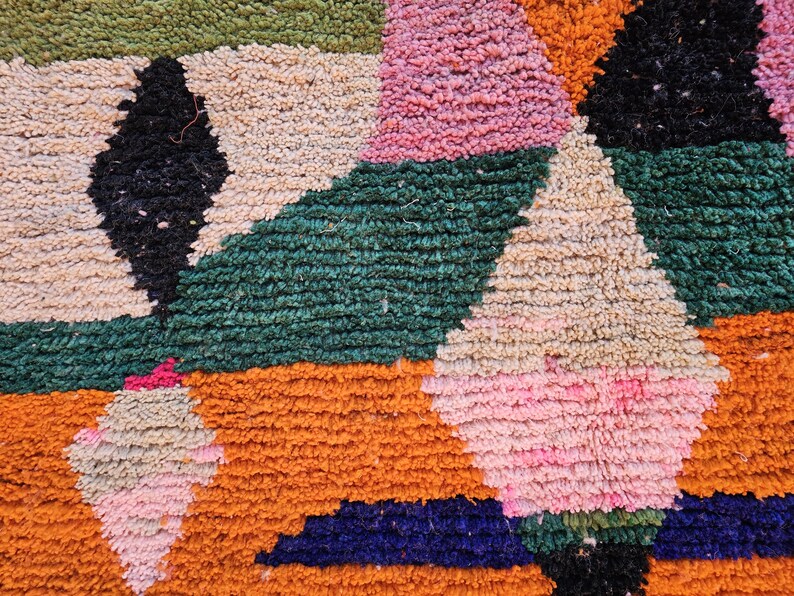 May include: Close-up of a colorful, textured rug with geometric patterns. The rug features a mix of colors including orange, green, pink, black, and cream. The design includes triangles and diamond shapes.