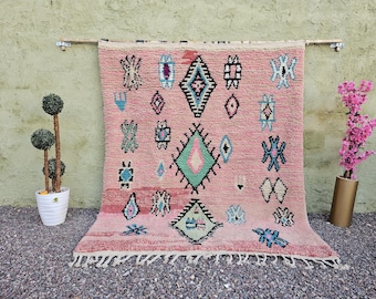 May include: A pink Moroccan rug with geometric patterns in black, teal, and white. The rug is rectangular with a fringe along the bottom edge. The rug is hanging on a wooden rod against a light green wall.