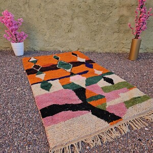 May include: A colorful geometric rug with a vibrant design. The rug features a mix of orange, pink, green, black, and white shapes. It has a fringed edge and is placed on a gravel surface. Decorative pink flowers in white and gold vases are in the background.