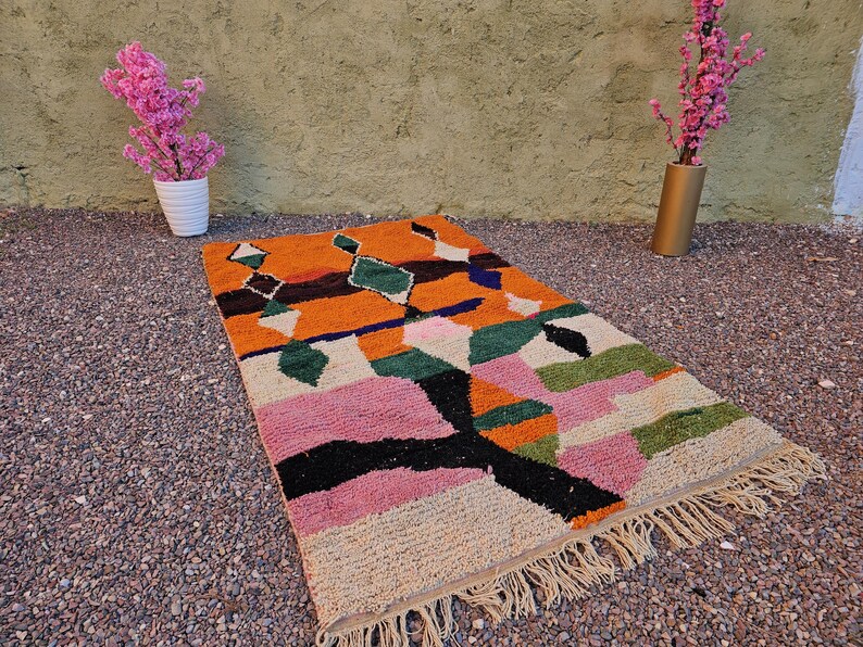 May include: A colorful geometric rug with a vibrant design. The rug features a mix of orange, pink, green, black, and white shapes. It has a fringed edge and is placed on a gravel surface. Decorative pink flowers in white and gold vases are in the background.