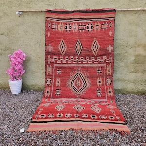 May include: A red and black patterned rug with geometric designs and fringe. The rug features diamond and rectangular shapes in a symmetrical pattern. The rug is displayed outdoors with pink flowers in pots on either side.