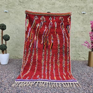 May include: A vibrant red rug with a textured surface and a pattern of vertical lines in various colors. The rug has a fringe along the bottom edge and is displayed against a neutral background. The rug is a decorative home decor item.