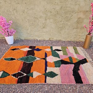 May include: A rectangular, hand-woven rug with a vibrant geometric design. The rug features an orange base with diamond and stripe patterns in green, black, white, pink, and blue. The rug is approximately 3 x 5 feet.