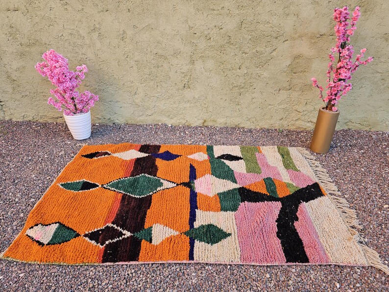 May include: A rectangular, hand-woven rug with a vibrant geometric design. The rug features an orange base with diamond and stripe patterns in green, black, white, pink, and blue. The rug is approximately 3 x 5 feet.
