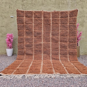 May include: A large, rectangular brown rug with a textured, grid-like pattern. The rug has a fringe along the bottom edge. The rug is displayed outdoors, leaning against a textured wall, with pink flowers in pots on either side.
