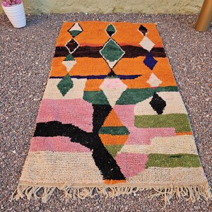 May include: A rectangular, colorful rug with a geometric design. The rug features a vibrant orange background with diamond and rectangular shapes in green, pink, black, and white. The rug has a fringed edge and is placed on a textured surface.
