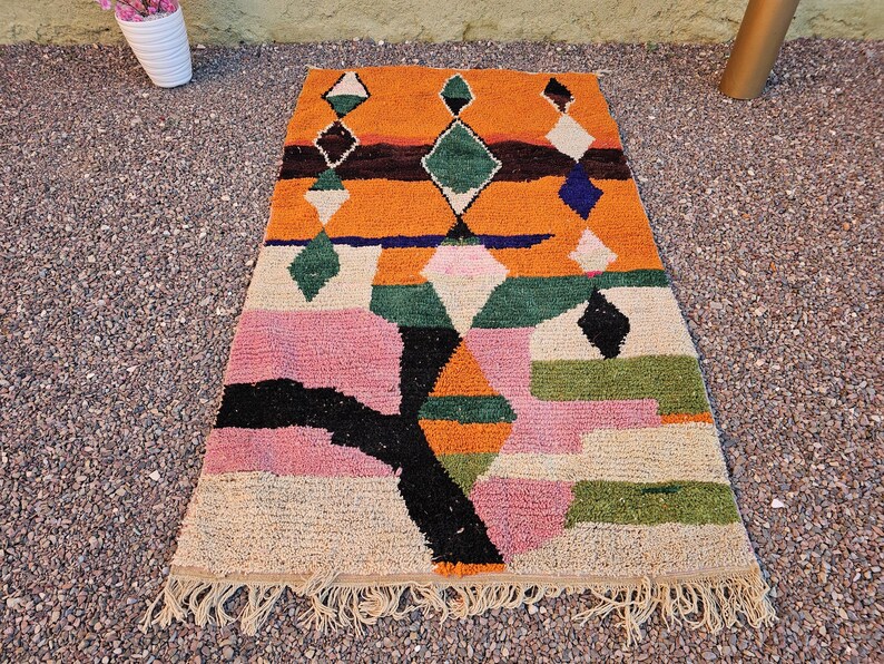 May include: A rectangular, colorful rug with a geometric design. The rug features a vibrant orange background with diamond and rectangular shapes in green, pink, black, and white. The rug has a fringed edge and is placed on a textured surface.