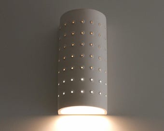 Puede incluir: Aplique de pared cilíndrico blanco con una serie de pequeños agujeros espaciados uniformemente. La lámpara emite una luz cálida, proyectando un suave resplandor sobre la pared de abajo.