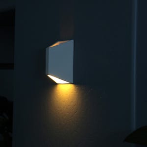 May include: A white, angular wall sconce with a warm, yellow light shining down on a textured wall.