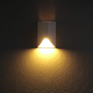 May include: A white, geometric wall sconce with a triangular light source. The sconce is mounted on a textured, dark brown wall. The light emits a warm, yellow glow, creating a contrast against the dark background. This fixture is designed for indoor lighting.