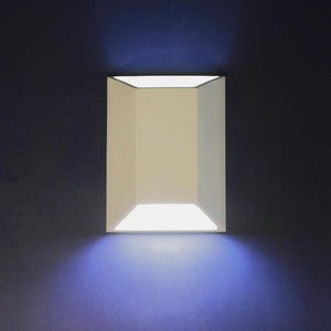 May include: A rectangular, cream-coloured wall sconce with a geometric design. The sconce emits a bright, white light, casting a blue glow on the dark blue wall. The fixture has a modern, minimalist aesthetic.