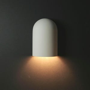 May include: A white, arched wall light emitting a warm glow from its base. The light is mounted on a dark grey wall, creating a contrast that accentuates the minimalist design of the sconce. This modern lighting fixture adds a touch of elegance to any space.