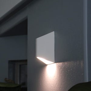 May include: A white wall-mounted light fitting with a rectangular shape and a slanted edge. The light is switched on and casts a warm glow on the wall.