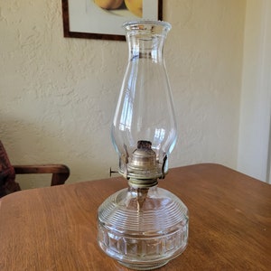 May include: A clear glass oil lamp with a brass burner and a textured glass base. The lamp is sitting on a wooden table.