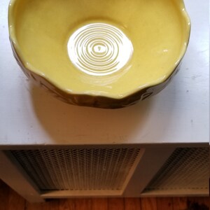 May include: A yellow ceramic bowl with a scalloped rim. The bowl has a glossy finish and a circular pattern in the center. The bowl is sitting on a white surface.