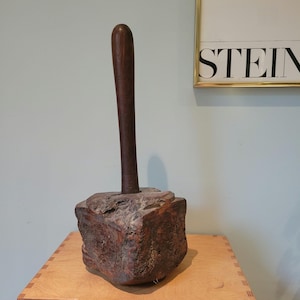 Puede incluir: Un mazo de madera grande con un mango grueso de color marrón oscuro. La cabeza del mazo tiene una forma irregular y rugosa y también está hecha de madera marrón oscuro.