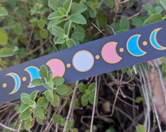 May include: A rectangular dark gray bookmark featuring a horizontal sequence of moon phases. The lunar cycle is depicted with gold outlines and filled with alternating light blue, pink, and white colors, representing the full progression of the moon.