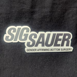 May include: A black surface with a white and gray "SIG Sauer" logo and the text "GENDER AFFIRMING BOTTOM SURGERY" in a white font.