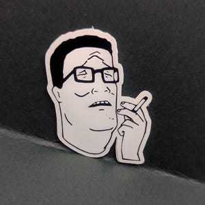 May include: A white sticker featuring a black and white cartoon illustration of a man with glasses, a furrowed brow, and a cigarette in his hand. The sticker has a white border and is set against a dark background.