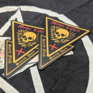 May include: Black and gold triangular stickers with the text "Medic Coven", a skull, and the words "Sacred Flesh" and "Sacred Duty". The stickers have a red cross symbol and are on a black and white patterned background.