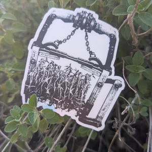 May include: A black and white sticker featuring a vintage bear trap design. The sticker shows a detailed illustration of the trap with chains and a wooden frame, set against a background of tall grass. The sticker is placed on a bed of green leaves.