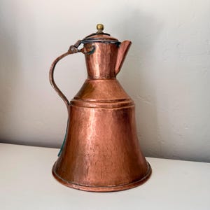 May include: A hammered copper jug with a curved handle and a small spout. The jug has a lid with a brass knob and a flared base. The copper has a rich, reddish-brown colour and a textured surface. The jug is likely vintage.
