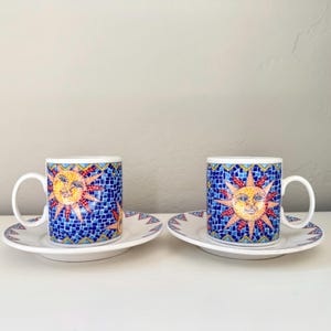 May include: Two white ceramic mugs and matching saucers featuring a vibrant blue mosaic pattern with a central yellow sun face design. The mugs have curved handles and the saucers display a geometric border pattern in blue, red, and yellow.