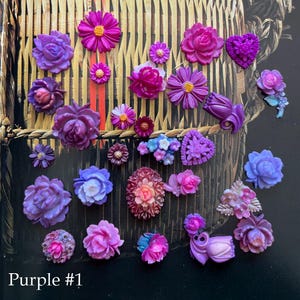 May include: A collection of purple flower-shaped embellishments in various sizes and styles. The assortment includes roses, daisies, and heart-shaped designs, with a few featuring blue and pink accents. The text "Purple #1" is visible at the bottom.