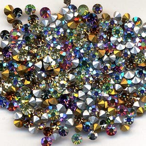 Puede incluir: Una colección de pedrería iridiscente en varios colores, incluyendo dorado, plateado, morado, verde y azul. Las piedras son redondas y facetadas, reflejando la luz. Ideal para manualidades y adornos.