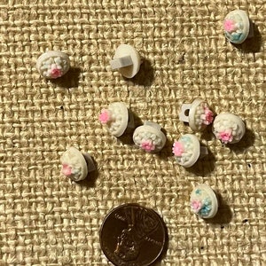 Puede incluir: Un conjunto de pequeños botones blancos con forma de flor, con detalles en rosa y azul. Cada botón tiene un tallo gris en la parte posterior. Se incluye una moneda como referencia de tamaño. Los botones están sobre una superficie beige texturizada.
