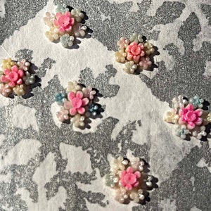 May include: Five small, pink and white flower embellishments with pearl-like accents. The embellishments are arranged in a scattered pattern on a white, textured surface.