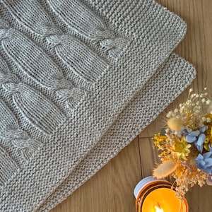 Op de afbeelding: Een lichtgrijze gebreide deken met een kabelbreipatroon. De deken is opgevouwen en toont de ingewikkelde textuur en het patroon. Een klein boeket droogbloemen en een brandende kaars staan op de achtergrond.