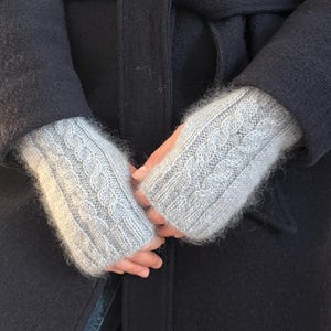 Puede incluir: Un par de guantes sin dedos grises con un patrón de punto de cable. Los guantes están hechos de un material suave y peludo.