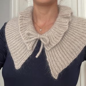 Puede incluir: Un cuello de punto desmontable en color beige con textura acanalada, borde con volantes y un pequeño lazo de cierre en el centro. El accesorio está confeccionado con hilo suave y esponjoso, sobre una camiseta azul marino de manga larga.