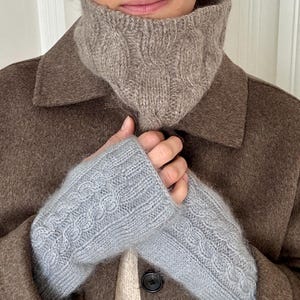 Puede incluir: Un calentador de cuello de punto trenzado beige y un par de guantes sin dedos de punto trenzado azul claro. El calentador de cuello presenta un patrón de trenzas verticales texturizado, mientras que los guantes muestran un diseño a juego.