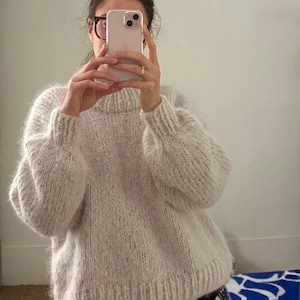 Peut inclure: Un pull en tricot épais de couleur avoine clair avec un aspect duveteux et texturé. Le pull présente un col rond côtelé, des manches longues avec des poignets côtelés et un ourlet côtelé. La maille est lâche et volumineuse.