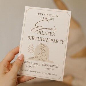Close-up of a hand holding a minimalist Pilates birthday invitation in a serene yoga studio setting. The beige and brown tones match the calm, self-care vibe, with a blurred background showing a woman stretching on a mat. Perfect for Etsy listings promoting wellness or Pilates-themed parties