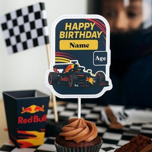 May include: A birthday cupcake with chocolate frosting, a cake slice, and a Red Bull-themed party decoration. The centerpiece is a cake topper with a race car and the words "Happy Birthday" and space for a name and age. A checkered flag is in the background.