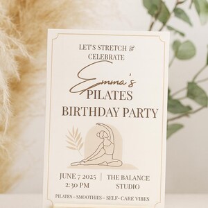 Minimalist Pilates birthday invitation displayed upright in a light wooden holder, surrounded by pampas grass and eucalyptus. Neutral beige background with soft natural lighting, highlighting the elegant boho design. Perfect for wellness or self-care themed parties.