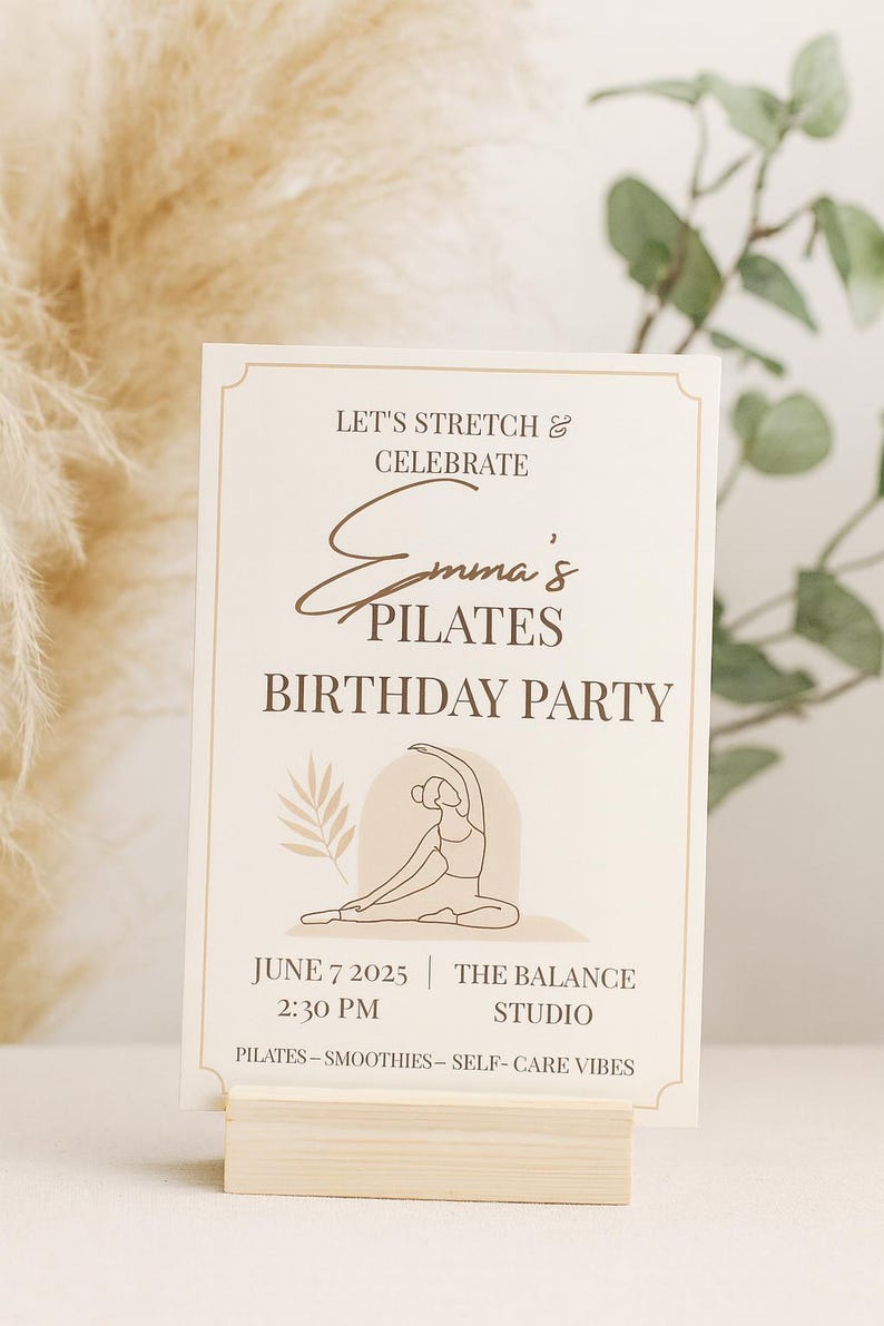 Minimalist Pilates birthday invitation displayed upright in a light wooden holder, surrounded by pampas grass and eucalyptus. Neutral beige background with soft natural lighting, highlighting the elegant boho design. Perfect for wellness or self-care themed parties.