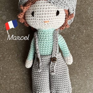 Peut inclure: Poupée au crochet faite à la main nommée "Marcel", vêtue d'une chemise bleu clair, d'une salopette grise avec des boutons en bois et d'un bonnet gris. La poupée a les cheveux et les pieds bruns. Un petit drapeau français est à côté de la poupée.