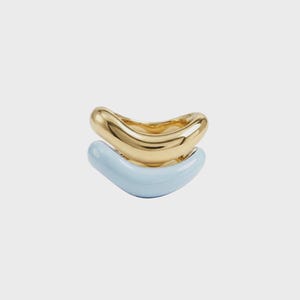 May include: Two stacked rings. The top ring is gold-colored, and the bottom ring is light blue. Both rings have a smooth, curved design. The rings are set against a plain, light background.