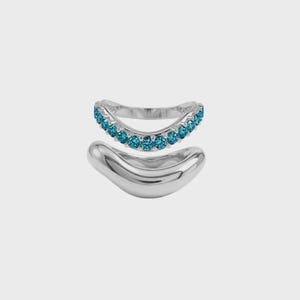 Puede incluir: Dos anillos de plata. El anillo superior es curvo y está engastado con pequeñas piedras redondas de color turquesa. El anillo inferior es una banda de plata lisa y maciza con una forma orgánica y fluida. Ambos anillos se muestran sobre un fondo blanco.
