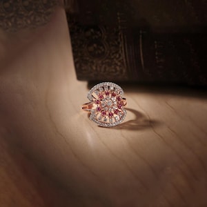 May include: Rose gold ring with a floral design. The ring features a central cluster of red gemstones surrounded by smaller white stones. The ring's setting has an openwork design, and it sits on a wooden surface.