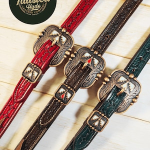 May include: Close-up of three leather belts in red, brown, and green. Each belt features a decorative buckle with a southwestern design and a matching smaller buckle. The belts are embossed with a detailed pattern and are displayed on a light wood surface.