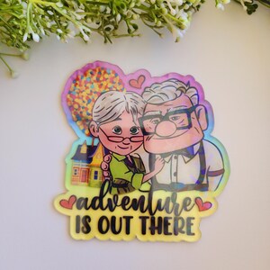 May include: A holographic sticker featuring an elderly couple, a colorful house with balloons, and a small heart. The text reads "adventure IS OUT THERE" in black script and block lettering. The sticker has a reflective, iridescent finish.