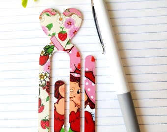 May include: A decorative paperclip featuring a heart and a cartoon character design with strawberries and floral patterns. A white and gray pen rests on a lined paper surface. The text "My Lovely Creations" is visible.